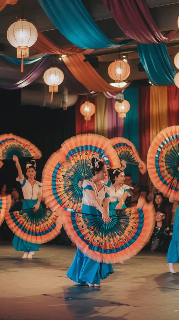A vibrant dance performance featuring dancers in colorful traditional attire holding large hand fans with bold peacock feather designs. The background is decorated with draped fabric in jewel tones and hanging lanterns, creating a festive ambiance.