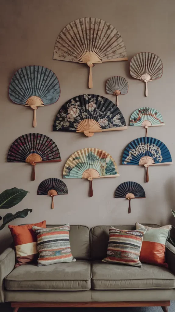 A cozy living room with a neutral-toned wall displaying an elegant arrangement of hand fans as wall art. The fans vary in size and design, featuring intricate floral patterns, traditional motifs, and bold colors. Below the display, a modern sofa with colorful patterned cushions complements the artistic decor. Two potted plants on either side add a touch of greenery to the setting.